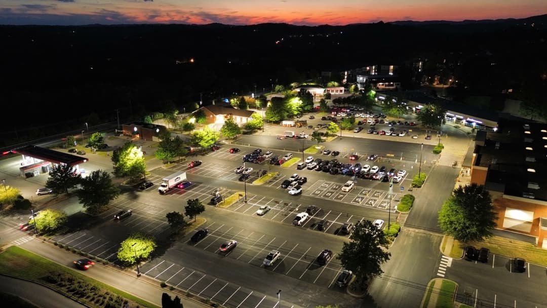 Aerial view of a partially filled parking lot at sunset, with illuminated businesses nearby.