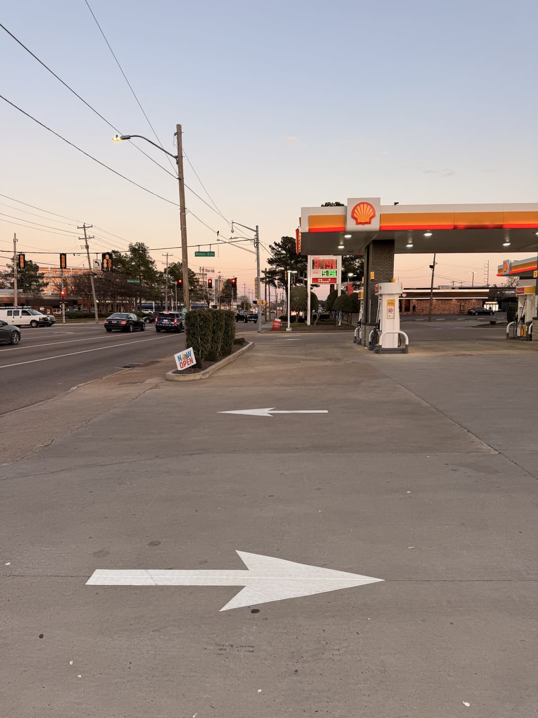 Shell gas station with directional arrows on pavement and street at dusk. Traffic signals visible.