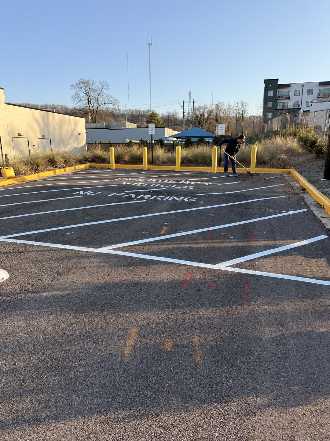 Man painting "No Parking" sign in empty parking lot with yellow bollards and surrounding buildings.